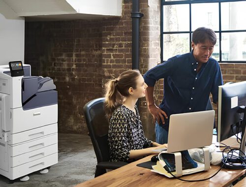 Un hombre y una mujer están uno al lado del otro frente a una computadora, concentrados en la pantalla. La impresora a color Xerox® VersaLink® C7000 está en el fondo.