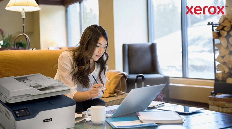Uma mulher trabalha em um laptop em um escritório confortável ou em uma sala de estar, com uma impressora Xerox compacta visível em primeiro plano, indicando integração perfeita da impressão com fluxos de trabalho digitais.