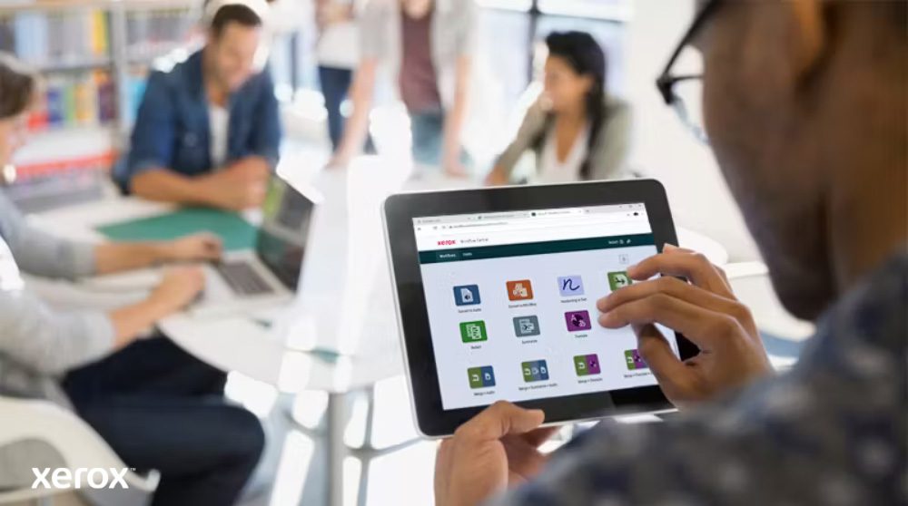 Plataforma Xerox® Workflow Central: acceso móvil intuitivo Una persona sostiene una tableta que muestra la interfaz de Xerox Workflow Central con varios íconos de aplicaciones, mientras otras personas trabajan en el fondo borroso, resaltando el acceso móvil a las soluciones de flujo de trabajo.