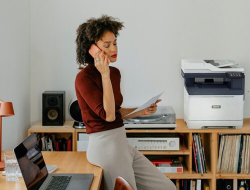 Uma mulher sentada em uma mesa recebendo uma ligação ao lado de uma impressora Xerox® C325