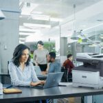 Mujer profesional escribiendo en una computadora portátil en un escritorio en un entorno de oficina junto a una impresora Xerox® C325