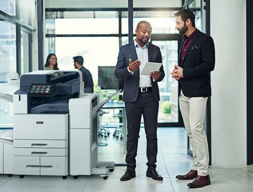 Zwei Männer in einem Büro diskutieren etwas auf einem Tablet neben einem Farbmultifunktionsdrucker der Xerox® AltaLink® C8200-Serie