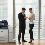 Duas pessoas num escritório em fatos de negócios a discutir documentos ao lado de uma impressora multifunções Xerox® AltaLink® série B8200