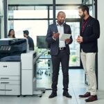 Dos hombres en una oficina conversan sobre algo en una tableta junto a una impresora multifunción en color Xerox® AltaLink® C8200 Series