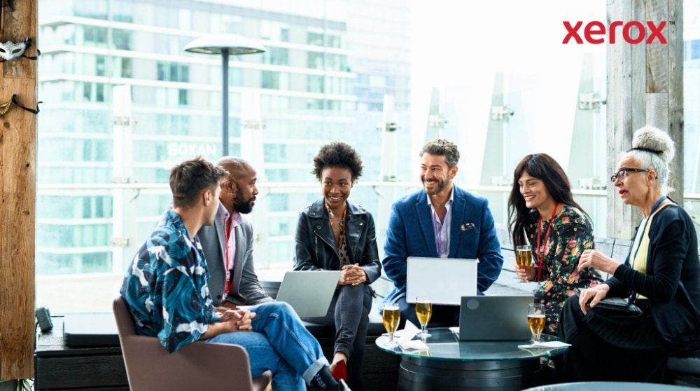 Um grupo de sete pessoas sentadas juntas, a conversar e a trabalhar em computadores portáteis com uma cidade ao fundo