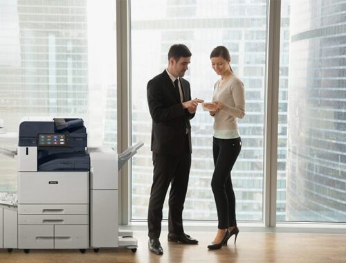 Two people in an office in business attire discussing documents stand by a Xerox® AltaLink® B8200 Series Multifunction Printer