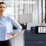 Eine Frau steht neben dem Xerox® PrimeLink® C9200 Serie Drucker in einer modernen Büroumgebung