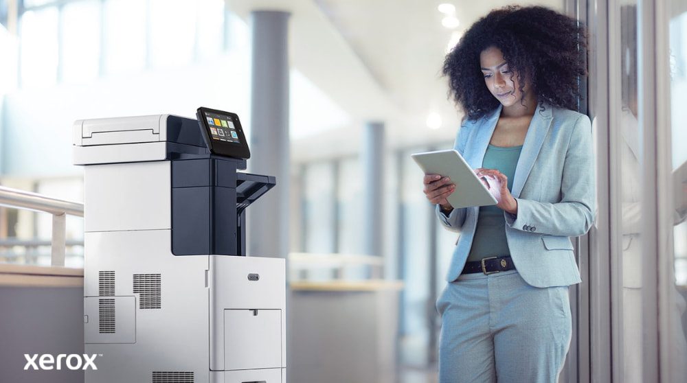 Uma pessoa está ao lado de uma impressora multifuncional Xerox VersaLink B605, olhando e interagindo com um tablet, ilustrando como o ConnectKey permite a interação móvel com a impressora.