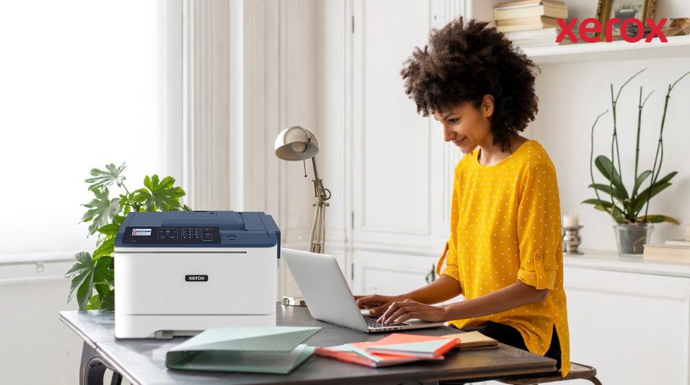 A person with curly hair is smiling while typing on a laptop at a desk, with a compact Xerox C310 printer positioned next to the laptop, showcasing a modern home office setup for productivity.
