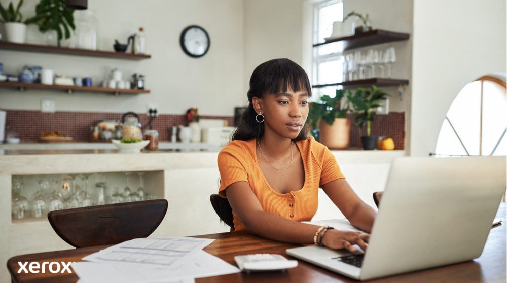 Une personne est assise à une table de cuisine, concentrée sur son travail sur un ordinateur portable, avec des papiers et une calculatrice à proximité, illustrant la flexibilité du travail à domicile avec une gestion efficace des documents.