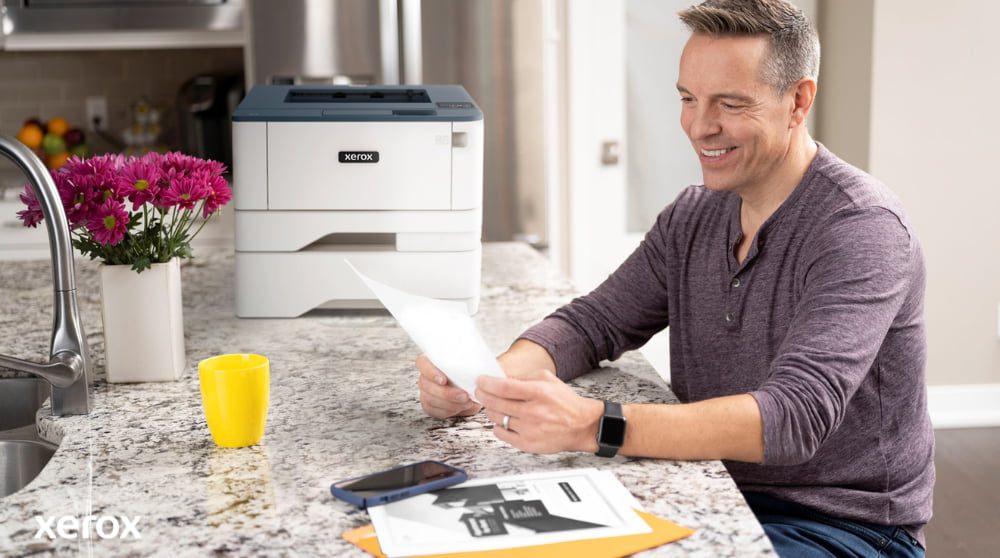 A person is smiling while reviewing a document at a kitchen counter, with a compact Xerox printer and a bright yellow mug nearby, suggesting seamless printing and document handling in a home or small office setting.