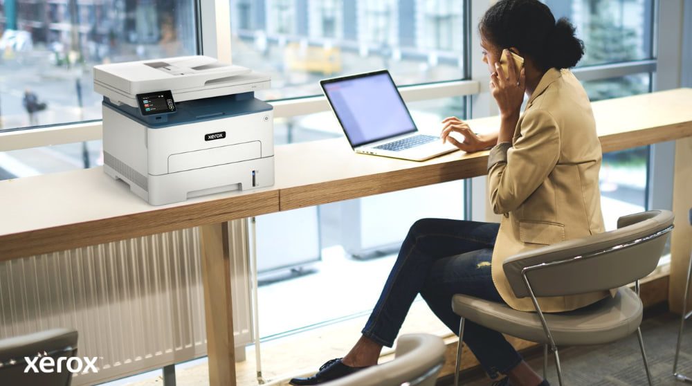 Una persona está sentada en una mesa alta junto a un gran ventanal, hablando por teléfono mientras trabaja con una laptop. Una impresora multifunción Xerox B235DNI está convenientemente colocada sobre la mesa junto a ella, lo que sugiere facilidad de uso en un espacio de trabajo moderno.