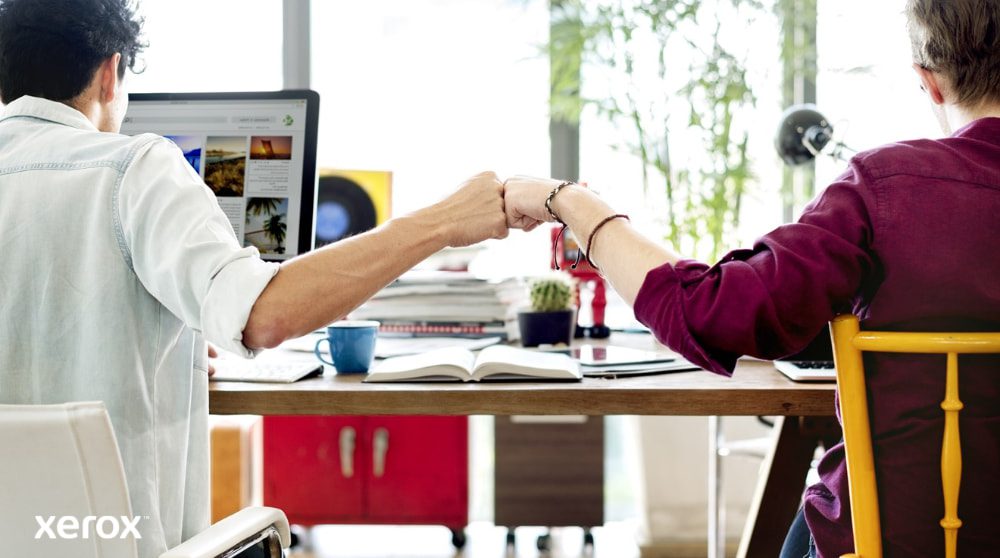 Xerox® Smart Start: Teamwork en samenwerking Twee mensen zitten aan een bureau, met hun gezicht van de camera af, en geven elkaar een 'fist bump', wat teamwork en succesvolle samenwerking symboliseert. De een kijkt naar een computerscherm.