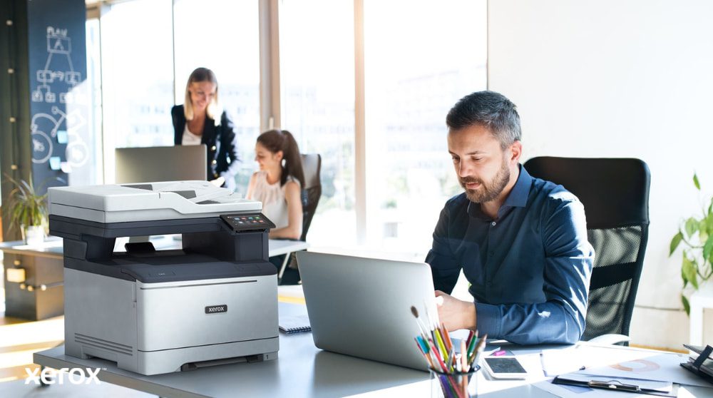 A person is seated at a desk, looking at a laptop with a Xerox C325 multifunction printer beside it. In the background, two other people are also working at their desks in a bright office environment.