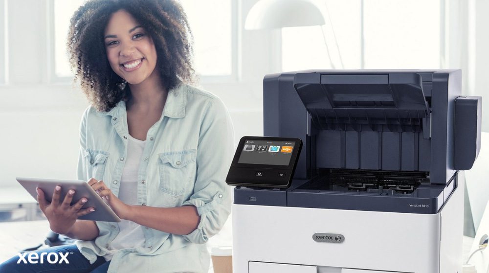 Xerox® Workplace Cloud e Suite: Integração fácil de usar Uma pessoa sorridente está sentada ao lado de uma impressora Xerox VersaLink B610 branca, segurando um tablet e olhando para a câmera, destacando a natureza amigável das soluções Xerox.